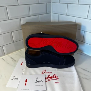 Mörkblå sneakers från Christian Louboutin - Snygga mörkblå sneakers från Christian Louboutin med klassisk röd sula och rund tå. Skorna har snörning och är tillverkade i mocka med broderad logga baktill. Perfekt för dig som vill sticka ut med exklusiv stil och detaljer.