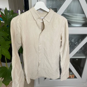 Beige linneskjorta Knowledge Cotton - Snygg beige skjorta från Knowledge Cotton Apparel i storlek S. Tillverkad i svalt linne med klassisk button down-krage och långa ärmar. Skjortan har en stilren look med knappar framtill och passar perfekt till jeans eller chinos.