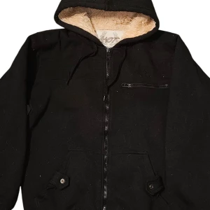 Svart teddyjacka från Hot Collection - Svart teddyjacka med mysigt beige foder, huva och dragkedja framtill. Jackan har en bröstficka med dragkedja och två fickor med knappdetaljer. Perfekt för kalla dagar när du vill hålla stilen och värmen.