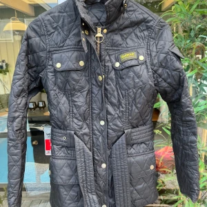 Svart quiltad jacka från Barbour - Quiltad svart jacka från Barbour med bälte i midjan, guldiga knappar och dragkedja. Klassisk design med fyra fickor framtill och hög krage. Perfekt för dig som gillar stilren och tidlös look. Jackan har Barbour-logga på bröstet och markerad midja.