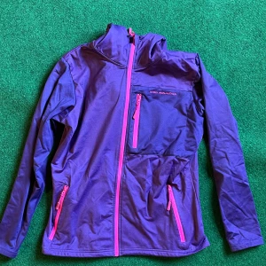 Lila vindjacka från High Mountain - Sportig lila vindjacka från High Mountain med rosa dragkedjor och flera fickor. Jackan har lång ärm, hög krage och är perfekt för utomhusaktiviteter. Snygg och funktionell design som sticker ut med sina färgdetaljer.