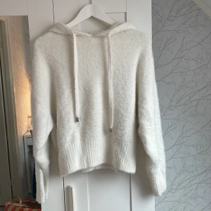 Vit fluffig hoodie från H&M - Supermjuk och fluffig vit hoodie från H&M i storlek S. Tröjan har huva med snörning och långa ärmar. Den är stickad och har en mysig känsla, perfekt för kyliga dagar. Ribbstickad nederkant och ärmslut ger en snygg finish.