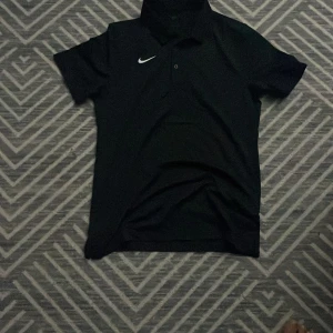 Svart pikétröja från Nike i S - Svart pikétröja från Nike med klassisk krage och korta ärmar. Tröjan har en vit Nike-logga på bröstet och är tillverkad i ett lätt och svalt polyestermaterial som passar perfekt för träning eller chill dagar.PRIS KAN DISKUTERAS 