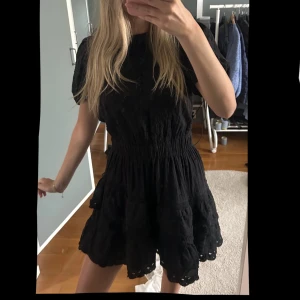 Svart kort klänning med volang - Supersöt svart kort klänning med volangkant och markerad midja. Klänningen har korta ärmar och en luftig, lätt struktur som ger en romantisk vibe. Perfekt för dig som gillar en feminin och trendig stil.