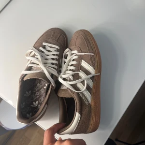 Adidas Speziales i mocka och beige bruna - Säljer ett par Adidas Samba sneakers i beige mocka med vita detaljer och klassiska tre ränder på sidan. Skorna har vit snörning, gummisula och rund tå. Perfekta för dig som gillar retrostil och vill ha något som sticker ut men ändå är lättmatchat.