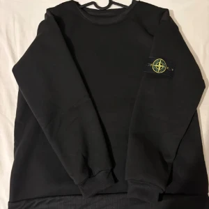 Svart Stone Island sweatshirt - Svart sweatshirt från Stone Island med klassisk patch på vänster ärm. Tröjan har rund halsringning, ribbade muddar och är tillverkad i mjuk bomull. Perfekt för dig som gillar stilrena och ikoniska streetwear-plagg.