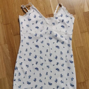 Vit blommig kortklänning från HM  - Supersöt vit kortklänning från Divided HM med blått blommönster, smala axelband och spetsdetaljer vid urringningen. Klänningen är ribbad och har en liten rosett framtill. Perfekt för sommarens varma dagar! Hör av er vid eventuella frågor💕💕