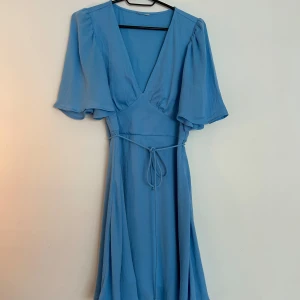 Blå omlottklänning med knytband - Snygg blå klänning med omlottmodell och v-ringning. Klänningen har vida, korta ärmar och ett smalt knytband i midjan som ger fin passform. Pris går att diskutera.