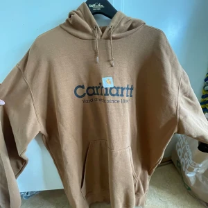 Brun Carhartt hoodie med tryck XL - Säljer en brun hoodie från Carhartt med stor logga och text på bröstet. Tröjan har huva med snörning och en stor magficka. Perfekt för dig som gillar streetwear och vill ha en chill look.