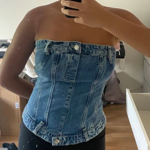 Jeans tubtopp från Zara - Säljer en snygg jeans och knappt använd tubtopp från Zara i klassisk blå denim. Toppen har bandeau-modell, bältdetaljer, knapp framtill och dragkedja bak. Sitter tight och har coola sömmar och fickdetaljer som ger en unik look. 