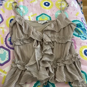 Beige volangtop från Amisu - Supersöt beige linnetopp från Amisu med tunna axelband, volanger och ribbad struktur. Toppen har omlottkänsla och knyt fram, vilket ger en romantisk vibe. Perfekt för dig som gillar detaljer och en luftig känsla.