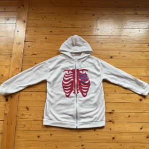 Vit hoodie med skelettmotiv XL - Vit hoodie med dragkedja och huva, prydd med ett stort rött och blått skelettmotiv på framsidan. Skön och avslappnad passform, perfekt för dig som gillar unika och lite edgy plagg. Tillverkad i mjuk bomullsmix. Står XL men är L