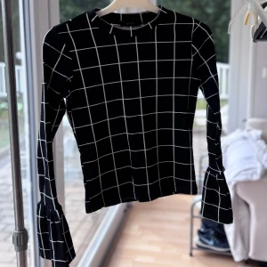 Svart rutig topp med trumpetärm  - Svart långärmad topp med vitt rutmönster och trendiga trumpetärmar. Sitter snyggt med normal passform och har en rund halsringning. 