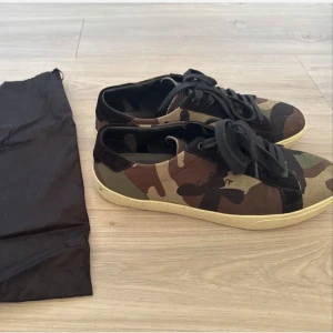 Saint lauren sneakers - Snygga sneakers från Saint Laurent i klassiskt camouflage-mönster med svarta detaljer och svarta skosnören. Yttermaterialet är canvas och mocka, sulan är beige. Perfekta för dig som vill sticka ut med en cool streetstyle.