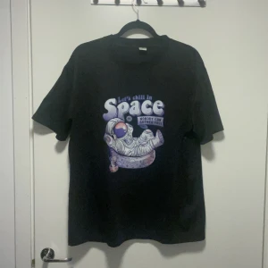Svart t-shirt med rymdtryck - Svart t-shirt med coolt tryck av en astronaut som chillar i rymden och texten 'Let's chill in Space, nobody can bother you!'. T-shirten har korta ärmar och avslappnad passform. Perfekt för dig som gillar unika och lekfulla motiv. Storlek L men skulle liten i storlek