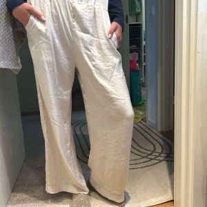 Vida ljusa byxor i satinlook - Säljer ett par superbekväma, vida byxor i ljus beige med snygg satinliknande finish. De har resår i midjan och djupa fickor framtill. Perfekta för dig som gillar en avslappnad men ändå stilren vibe. Silke material