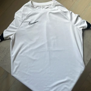 Vit Nike fotbollströja - Snygg vit fotbollströja från Nike med svart swoosh-logga på bröstet och svart rand på ärmarna. Tröjan är kortärmad och tillverkad i ett lätt och ventilerande material som passar perfekt för träning eller match. Oanvänd så 10/10 skick.