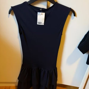 Marinblå volangklänning från H&M - Snygg marinblå klänning från H&M med volangdetalj nedtill. Ärmlös modell med rund halsringning och figurnära passform upptill. Perfekt för dig som gillar stilrena och trendiga plagg.