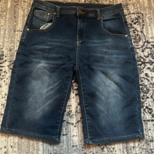 Mörkblå jeansshorts från Knox - Snygga mörkblå jeansshorts från Knox med klassisk femficksdesign och kontrastsömmar. Modellen är rak och slutar strax ovanför knäna. Perfekta för sommaren och har en cool urban vibe. Passar dig som gillar en enkel men stilren look.
