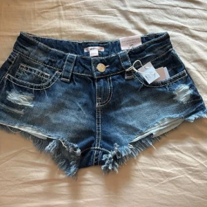 Blåa lågmidjade jeansshorts  - Snygga blå jeansshorts från Gina Tricot i låg midja med slitna detaljer och fransig kant. Klassisk femficksmodell med coola slitningar framtill och dekor på bakfickorna. Perfekta för varma sommardagar och festivalhäng.