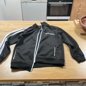 Palm Angels svart track jacket - Svart track jacket från Palm Angels med vita ränder längs ärmarna och vit dragkedja. Jackan har ståkrage, två fickor med dragkedja och logga broderad på bröstet. Perfekt för en sportig och trendig streetstyle.