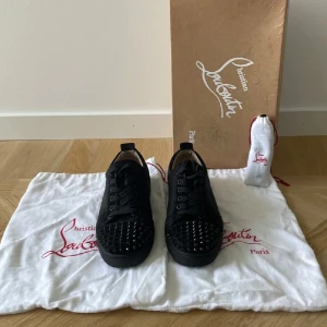 Christian Louboutin svarta sneakers med nitar - Svarta sneakers från Christian Louboutin med mattsvarta nitar på tån och klassisk röd sula. Skorna har snörning, rund tå och är tillverkade i skinn och mocka. Snygg och edgy design som sticker ut, perfekt för dig som gillar statement-detaljer.