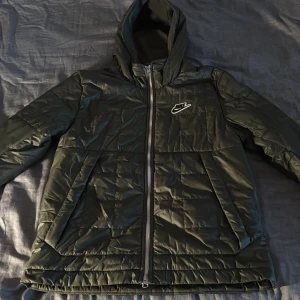 Mörkgrön pufferjacka från Nike - Säljer en mörkgrön pufferjacka från Nike med huva och hel dragkedja. Jackan har quiltad utsida och mjukt fleecefoder på insidan för extra värme. Klassisk Nike-logga på bröstet och två stora fickor framtill. Perfekt för kalla dagar och snygg streetstil.