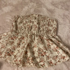 Blommig bandeau tubtopp i vitt och rosa - Supersöt bandeau tubtopp med smockad överdel och volangig nederdel. Vit botten med små blommor i rosa och grönt, perfekt för sommarens alla tillfällen. Sitter uppe utan axelband och har en luftig känsla.