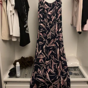 Mönstrad långklänning från Jyothi - Snygg långklänning från Jyothi med marinblå bas och rosa bladliknande mönster. Klänningen har smala axelband desig, perfekt för en somrig look. Lätt och flowy känsla som ger en avslappnad men ändå stilren vibe. Säljer då jag tycker om mer korta klänningar än långa och gör att denna inte kommer så mycket till användning. 