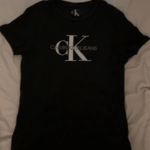 Svart t-shirt från Calvin Klein Jeans - Svart t-shirt från Calvin Klein med stort vitt CK-logotryck på bröstet. Snygg och enkel modell som passar perfekt till jeans eller kjol. Mjuk och skön till sommaren. Säljer då den inte passar mig längre. Sitter som både M och S. 