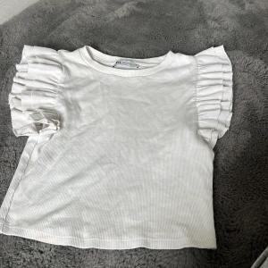 Vit volangärmad topp från Zara - Supersöt vit topp från Zara med korta ärmar och breda volangdetaljer på axlarna. Ribbad struktur i tyget ger en extra touch. Perfekt för dig som gillar en feminin och trendig stil.