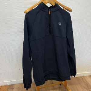 Svart Morris half zip hoodie med brodyr - Svart hoodie med half zip och broderad scoutlilja på bröstet. Tröjan har hög krage, långa ärmar och är gjord i mjuk bomull. Perfekt för chill dagar eller när du vill ha en enkel men snygg stil.