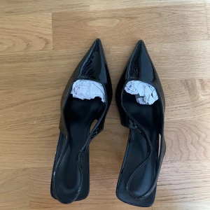 Svarta lackade pumps med spetsig tå - Snygga svarta pumps i lack med spetsig tå och öppen häl. Skorna har en smal klack och elegant slingback-design som ger en stilren och trendig look. Perfekta för dig som vill sticka ut med klassiska men edgy klackar. Helt nya. Personen på bilden har storlek 38.