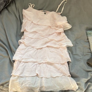 Ljusrosa volangklänning med smala axelband - Söt ljusrosa kortklänning med flera lager volanger och smala axelband. Klänningen har en luftig känsla och är perfekt för varma dagar. Den är ärmlös och har en feminin, flowy siluett som ger en romantisk vibe.