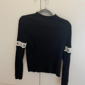 Svart stickad tröja Calvin Klein Jeans - Svart stickad tröja från Calvin Klein Jeans med vita band och CK-logga runt ärmarna. Tröjan har lång ärm, ribbad struktur och en klassisk passform. Perfekt för dig som gillar stilrena och trendiga plagg med tydlig logga. Kan mötas upp i Trollhättan eller Alingsås. Kan skickas men då tillkommer en fraktkostnad.
