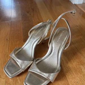 Guldiga sandaler med klack från Stradivarius - Snygga guldiga sandaler från Stradivarius med fyrkantig tå och smala remmar runt vristen. Skorna har en elegant låg klack och är tillverkade i syntetmaterial med metallic-finish. Perfekta för att lyfta vilken outfit som helst.