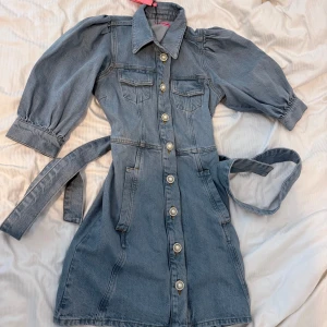 Jeansklänning med pärlknappar Custommade - Helt ny, Superfin jeansklänning från Custommade i ljusblå denim med puffärmar, pärlknappar och bälte i midjan. Klänningen har krage, fickor fram och trekvartsärm. Perfekt för dig som vill ha en cool och trendig look. Säljer eftersom jag köpte den som present till någon och kunde ej ge den! Kostar egentligen 3 099- säljer för 1000!!