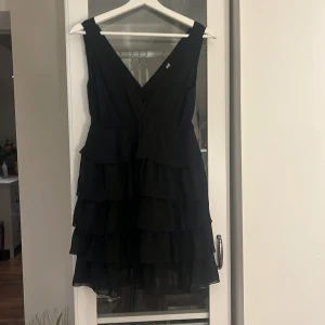 Svart volangklänning från H&M - Supersnygg svart kortklänning från H&M med flera lager volanger och v-ringad omlott framtill. Ärmlös modell i lätt tyg som ger en riktigt festlig känsla. Perfekt för dig som vill sticka ut med en klassisk men trendig look. 