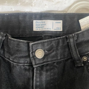 Svarta high waist straight jeans Bershka - Snygga svarta jeans från Bershka med high waist och raka ben. Klassisk femficksmodell i denim med knäppning fram och bälteshällor. Perfekta till sneakers eller boots för en clean look.
