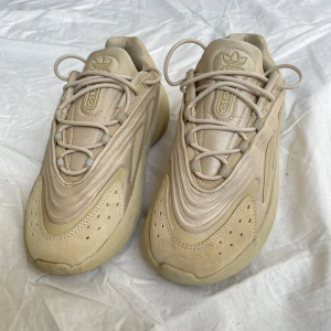 Beige Adidas Ozelia sneakers - Snygga Adidas Ozelia sneakers i beige med chunky sula och futuristisk design. Skorna har en mix av syntet och mocka, med klassiska 3-stripes detaljer på sidorna och snörning framtill. Perfekta för dig som gillar streetstyle. Endast använda 2 gånger. Nypris 1500. 