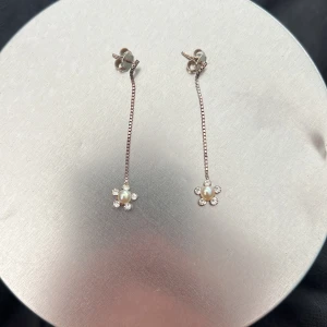 Silverörhängen med pärlblommor - Snygga silverörhängen med hängande kedja och blommor längst ner. Blommorna har en pärla i mitten och små glittrande stenar runt om. Perfekta för dig som gillar en söt och elegant stil med lite extra bling.