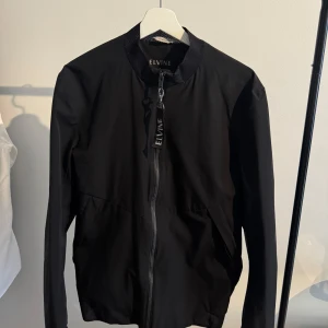 Svart vindjacka från Elvine - Snygg svart vindjacka från Elvine med dragkedja framtill och diskreta fickor. Jackan har en klassisk bomberkrage och stilren design, perfekt för dig som gillar minimalistiskt mode. Långärmad och passar till många olika outfits samt träning. 
