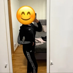 Svart EA7 mjukis set  med logga - Svarta mjukis set från EA7 Emporio Armani med vita loggor längs benen. Byxorna har en normal passform och resår i midjan. Byxorna är i storlek M och storlek S i koftan. 