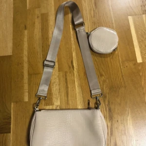 Beige axelväska med croc-mönster - Snygg beige axelväska i syntetmaterial med croc-mönster och bred justerbar axelrem. Väskan har en rund liten coin purse som sitter fast på remmen och stängs med dragkedja. Perfekt för att bära det viktigaste och ge outfiten en trendig touch. För mer bilder skriv till mig 