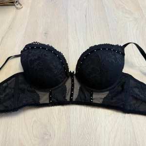 Svart spetskorsett från Censored - Supersnygg svart korsett från Censored med spetsdetaljer och små pärlor längs kuporna. Axelbandslös modell med transparent mesh och blommönstrad spets. Perfekt för dig som vill ha något extra till outfiten.