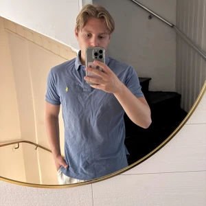 Blå pikétröja från Ralph Lauren - Säljer en klassisk blå pikétröja från Ralph Lauren med gul broderad logga på bröstet. Tröjan har krage, korta ärmar och är tillverkad i mjuk bomull. Perfekt för en clean och stilren look.
