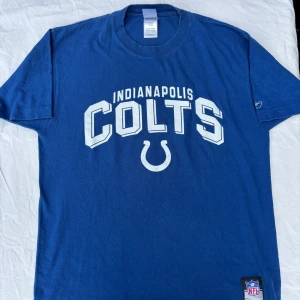 Blå Indianapolis Colts t-shirt Reebok - Blå t-shirt från Reebok med Indianapolis Colts-tryck i vitt framtill och NFL-märke nertill. Klassisk passform, rund hals och korta ärmar. Perfekt för dig som gillar amerikansk fotboll och vill ha en sportig look.