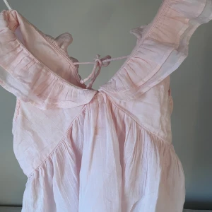 Ljusrosa volangtopp från  Gina Tricot - Supersöt ljusrosa klänning från Gina Tricot med volangdetaljer och knytning i ryggen. Klänningen är gjord i ett tunt, luftigt bomullstyg och har en romantisk känsla med sina rynkade partier och volanger längs axlarna. Perfekt för varma dagar och somriga vibes.