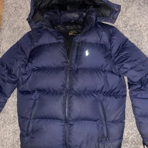 Mörkblå pufferjacka från Ralph Lauren - Säljer en snygg mörkblå pufferjacka från Ralph Lauren med dragkedja och huva. Jackan har en broderad logga på bröstet och är perfekt för kyliga dagar. Den har långa ärmar och två fickor med dragkedja framtill.