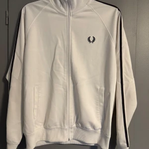 Vit track jacket från Fred Perry - Säljer en stilren vit track jacket från Fred Perry med svarta ränder längs ärmarna. Jackan har dragkedja framtill och ribbade muddar vid ärmslut och nederkant. Perfekt för en sportig look!